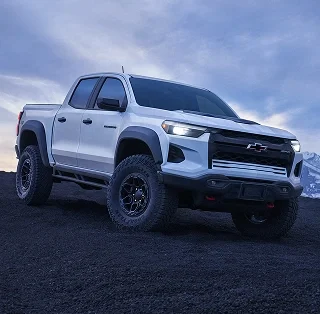 A white Chevrolet Colorado pickup with its headlamps on, parked on dark ground at dusk
