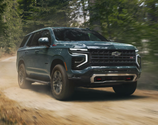 A dark teal Chevrolet Tahoe driving down a dirt road through a forest