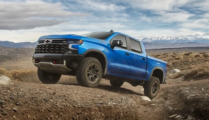 A blue Chevrolet Silverado 1500 climbing a rocky trail with snow-capped mountains behind it