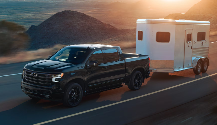 A black Chevrolet Silverado 1500 towing a white horse trailer along a desert highway at sunset